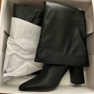 Boots från Nelly - Säljer ett par svarta höga stövlar i skinnimitation med bred klack och spetsig tå. Stövlarna har en stilren och minimalistisk look. De har en slät yta och går upp till knäet. Storlek 36 