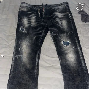 Svarta slitna jeans från märket dsquared2 - Säljer ett par svarta jeans med coola slitningar och patchade hål på benen. Jeansen har en tvättad look med ljusa partier och färgstänk, klassisk femficksmodell och dragkedja. Perfekta för dig som gillar en edgy streetstil. Skriv pm pm du undrar något