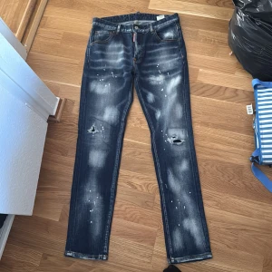 Mörkblå distressed jeans med färgstänk - Snygga mörkblå jeans med slitna detaljer och hål på knäna. Jeansen har coola vita färgstänk och en tvättad look som ger en edgy vibe. Klassisk femficksmodell med raka ben och kontrastsömmar. Perfekta för dig som gillar streetstyle.