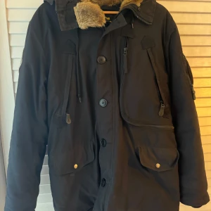 Svart parkajacka Alpha Industries M - Parkajacka från Alpha Industries i svart med huva och fuskpäls vid kragen. Jackan har flera fickor med både knappar och dragkedjor, samt patch med amerikansk flagga på ärmen. Perfekt för kalla dagar och har en cool militärvibe.