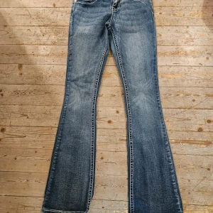 Blå bootcut jeans Fire & Rain - Snygga blå bootcut jeans från Fire & Rain med slitningar och markerade sömmar. Jeansen har coola bakfickor med lock och silvriga knappar, samt klassisk femficksdesign. Perfekta för dig som gillar en retro vibe och vill sticka ut lite extra.