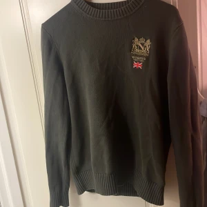 Mörkgrön  stickad tröja från Morris - Snygg mörkgrön stickad tröja från Morris med broderad logga och brittisk flagga på bröstet. Tröjan har rund halsringning och ribbade muddar vid ärmslut och nederkant. Perfekt för dig som gillar klassisk stil med en twist. Pris kan diskuteras.