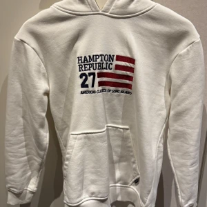 Vit hoodie från Hampton Republic - Vit hoodie från Hampton Republic med broderad logga och amerikanskt flaggmotiv på bröstet. Mjuk insida, stor huva och klassisk känguruficka framtill. Använd 1 gång, ny skick!