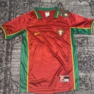 Vintage Portugal jersey  - Snabb leverans! 🚚. Dm för fler bilder!. Helt ny med tag! Säljer en vintage Portugal fotbollströja från Nike med Ronaldo och nummer 17 på ryggen. Tröjan är vinröd med gröna och gula detaljer, korta ärmar och klassisk krage. Materialet är lätt och andas, perfekt för match eller träning. Portugal-emblem på bröstet och Nike-logga.