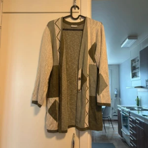 Grön och beige kofta med mönster - Snygg kofta i grönt och beige med geometriska mönster och två knappar framtill. Koftan har trekvartsärmar och stora fickor, perfekt för lager-på-lager-stil. Mjuk och skön, passar till chill outfits.