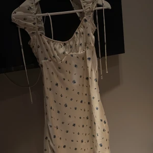 Blommig kortklänning från Zara - Supersöt kortklänning från Zara i ljus, nästan champagnefärgad ton med små blå blommor. Klänningen har puffiga korta ärmar med knytband och en lätt glansig yta. Perfekt för sommarens alla tillfällen och ger en romantisk vibe.