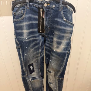Blå jeans med slitningar och dragkedja - Säljer ett par blå jeans med coola slitningar, patchdetaljer och färgstänk. Byxorna har en stor synlig dragkedja framtill och en avslappnad passform. Perfekta för dig som gillar en edgy streetstil.