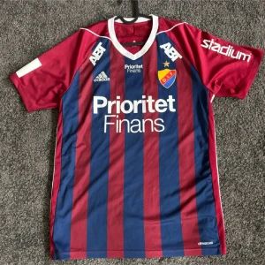 Djurgården fotbollströja Adidas - Snygg Djurgården fotbollströja från Adidas med breda blå och vinröda ränder, vita detaljer och klubbmärke på bröstet. Kortärmad modell i lätt och ventilerande material, perfekt för match eller träning. Sponsorer tryckta på bröst och ärmar.