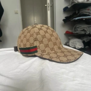 Gucci beige keps med GG-mönster - Säljer en beige keps från Gucci med klassiskt GG-monogram över hela kepsen och en snygg grön-röd rand på sidan. Justerbar rem baktill i brunt skinn. Materialet är bomull och kepsen har en rundad form med böjd skärm. Perfekt för dig som gillar lyxiga accessoarer.
