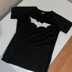 Svart Batman t-shirt i polyester - Compression - Svart t-shirt från Golden Vester med stor vit Batman-logga på bröstet. Tillverkad i mjuk och lätt polyester, perfekt för dig som gillar superhjältar och vill sticka ut med en cool print. Klassisk rund hals och korta ärmar. Passar även som XS