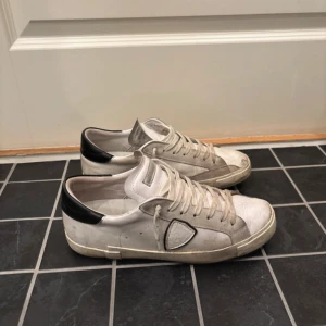 Philippe Model vita sneakers med svart häl - Snygga vita sneakers från Philippe Model med svart häl och klassisk logga på sidan. Skorna har rund tå, snörning och platt sula. Tillverkade i skinn och mocka för en cool och lyxig vibe. Perfekta för dig som gillar stilrena och trendiga sneakers.