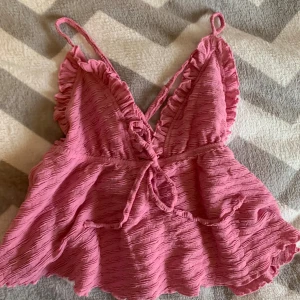 Rosa volangig linnetopp med knyt - Supersöt rosa linnetopp med volangkant och smala axelband. Toppen har en v-ringning med knyt framtill och är gjord i ett vågigt, strukturerat tyg som ger en cool look. Perfekt för varma dagar när du vill känna dig extra söt och somrig.