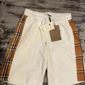 Vita Burberry shorts - Snygga vita shorts från Burberry med klassiska rutiga paneler i beige, svart och rött längs sidorna. De har elastisk midja med snörning för skön passform och är perfekta för sommaren. Stilen är clean men ändå lyxig, och materialet känns mjukt och bekvämt.