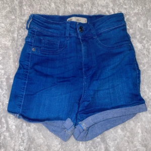 Blå högmidjade jeansshorts Molly XS - Snygga blå jeansshorts från Gina tricot  i storlek XS. Modellen är högmidjad med klassiska fem fickor, uppvikta benslut och bälteshällor. Perfekta för varma dagar när du vill ha en chill och trendig look.
