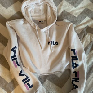 Vit croppad hoodie från FILA - Säljer en vit croppad hoodie från FILA med tryck på bröstet och båda ärmarna i marinblått och lila. Tröjan har huva med snörning och är tillverkad i mjuk bomull. Perfekt för dig som gillar sportig och chill stil.