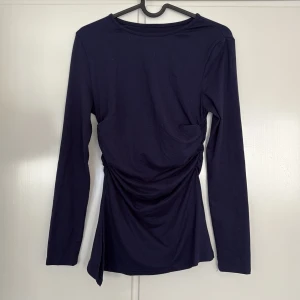 Mörkblå långärmad topp med drapering - Snygg mörkblå långärmad topp med draperad detalj i midjan som ger en cool och modern look. Toppen har rund halsringning och är tillverkad i mjukt bomullsmaterial. Perfekt att styla med jeans eller kjol för en trendig outfit.