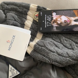Grå stickad Moncler hoodie med dragkedja - Säljer en grå stickad hoodie från Moncler med kabelstickat mönster och vita ränder på ena ärmen. Tröjan har quiltade partier på sidorna, dragkedja framtill och hög krage. Perfekt för kyliga dagar och snygg streetstil.