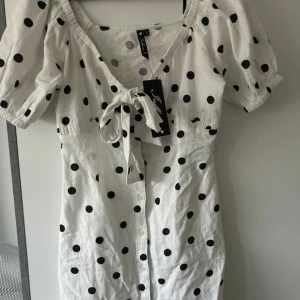 Vit prickig kortklänning Influence - Supersöt vit kortklänning från Influence med svarta prickar, puffärmar och knyt framtill. Klänningen har knappar längs framsidan och en feminin v-ringning. Perfekt för dig som gillar lekfulla och trendiga detaljer.