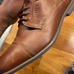 Bruna skinnloafers från AM Shoe Company - Snygga bruna loafers från AM Shoe Company i skinn med perforerade detaljer och kontrastsömmar. Skorna har snörning och en klassisk, stilren design med rund tå och platt sula. Perfekta för dig som vill ha en dressad look med en modern touch.