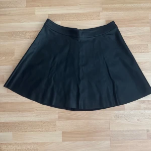 Svart kortkjol i skinnimitation - Snygg svart kortkjol i skinnimitation med lätt utställd form. Kjolen har en silvrig dragkedja bak som detalj och en stilren look som passar till många olika outfits. Perfekt för dig som gillar en edgy vibe. Använd en gång. Storleklapp avklippt, midjan 83cm, längd 42cm.