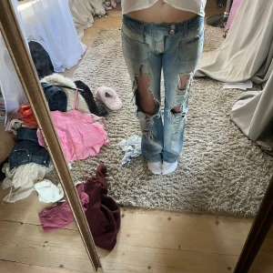 Ljusblå slitna jeans med raka ben - Säljer ett par ljusblå jeans med stora slitningar och hål på framsidan av benen. Jeansen har raka ben, klassiska fickor och låg midja. Perfekta för en avslappnad och trendig look.