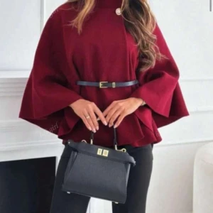 Vinröd cape med bälte och knapp - Stilren vinröd kappa med svart bälte i midjan och dekorativ guld knapp vid halsen. Jackan har en lös, svepande passform och hög krage. Perfekt för dig som vill ha en elegant och trendig look under höst och vinter.