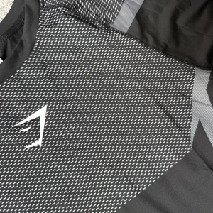 Svart Gymshark t-shirt  - Säljer en svart och grå Gymshark t-shirt i storlek M med korta ärmar och sportigt mönster framtill och baktill. T-shirten har ett diskret tryckt Gymshark-logo på bröstet och är gjord i ett lätt och stretchigt polyestermaterial som andas. Perfekt för dig som gillar att träna och vill ha en snygg look.