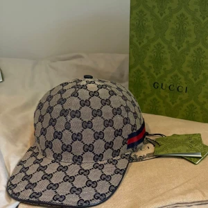 Gucci keps med GG-mönster - Snygg keps från Gucci i beige och mörkblått med klassiskt GG-monogram över hela kepsen. Kepsen har en böjd skärm och en röd-blå rand på sidan. Tillverkad i tåligt material med exklusiv känsla, perfekt för dig som vill sticka ut.