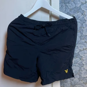 Svarta shorts från Lyle & Scott XS - Svarta bad  shorts från Lyle & Scott i storlek XS. Den har den ikoniska gula loggan på vänstra låret och sen en ficka på baksidan. Perfekt för dig som vill ha ett par snygga badshorts till stranden.