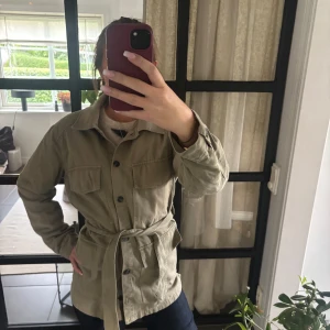 Grön jacka med bälte - Snygg olivgrön jacka med knappar framtill, två bröstfickor och knytskärp i midjan. Jackan har klassisk krage och är tillverkad i bomull, perfekt för lager-på-lager. Passformen är normal och ärmarna är långa. Enkel och stilren look som funkar till det mesta.