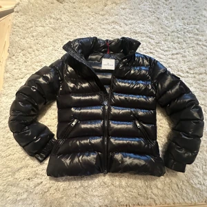  Svart Moncler pufferjacka  - Säljer en svart, glansig pufferjacka från Moncler med klassisk logga på ärmen. Jackan har dragkedja framtill, två snedställda fickor med dragkedja och hög krage. Insidan har ett coolt serietidningsmärke. Perfekt för kalla dagar och riktigt snygg streetstil. Byter hälst mot någon annan jacka typ canda gosse eller moncler ge förslag grejen man hänger i är trasig 