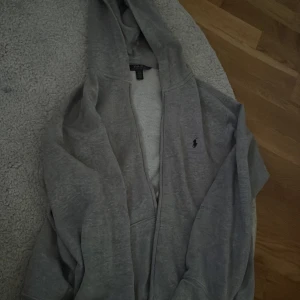 Grå hoodie från Polo Ralph Lauren - Säljer en grå hoodie från Polo Ralph Lauren med klassisk broderad logga på bröstet. Tröjan har huva, dragkedja framtill och är tillverkad i mjuk bomullsmix. Perfekt för chill dagar eller när du vill ha en clean och enkel look. Sälja för 599kr