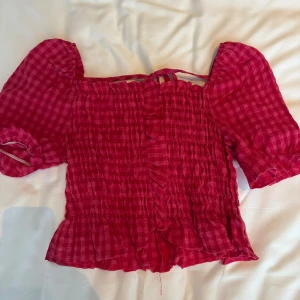 Rutig rosa blus med puffärm och volang - Supersöt rutig blus i stark rosa med puffiga korta ärmar, smockad kropp och volangkant nertill. Blusen har knytband upptill och en luftig, feminin känsla. Perfekt för dig som gillar romantiska detaljer och trendig stil.