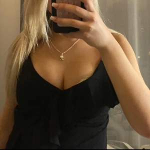 Svart omlott-linnetopp med volang - Säljer en svart linne-topp med omlottkänsla och volangdetalj framtill. Toppen har smala axelband och djup v-ringning, vilket ger en snygg och trendig look. Perfekt att styla med jeans eller kjol för en cool outfit.