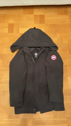 Svart Canada Goose  - Svart parkas från Canada Goose och äkta pälsdetalj. Jackan har klassisk röd logotyp på ärmen, dragkedja och flera fickor. Perfekt för kalla dagar med sin dunfyllda insida och robusta design.