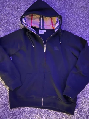 Svart Burberry hoodie - Svart hoodie från Burberry i storlek M med klassisk rutig insida på huvan och snygga snören. Tröjan har dragkedja framtill, fickor och ribbade muddar. Perfekt för dig som vill ha en stilren och lyxig look med ikonisk Burberry-detalj.