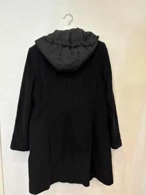 Svart kappa med huva från KappAhl - Svart kappa från KappAhl med snygg huva och dragkedja framtill. Jackan har långa ärmar, två fickor och en stilren look. Yttermaterialet är ullblandning och fodret är i syntet. Perfekt för dig som vill ha en klassisk och varm jacka med modern touch.