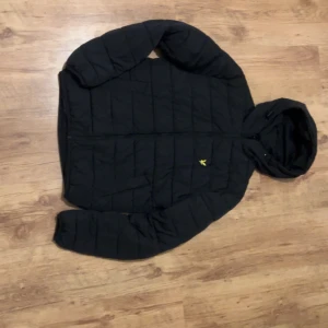 Svart pufferjacka från Lyle & Scott - Svart pufferjacka från Lyle & Scott med gul broderad logga på bröstet. Jackan har huva, dragkedja och quiltad design som ger en schysst look. Perfekt för kalla dagar och har långärmade ärmar. Snygg och enkel att matcha med det mesta.