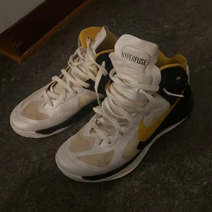 Nike Hyperfuse basket skor vit/gul/svart - Nike Hyperfuse sneakers med högt skaft i vit, gul och svart. Snyggt designade med gul swoosh och detaljer, meshpaneler för ventilation och robust snörning. Skön innersula och slitstark yttersula med mönster för extra grepp. Perfekta för dig som gillar sportig stil.