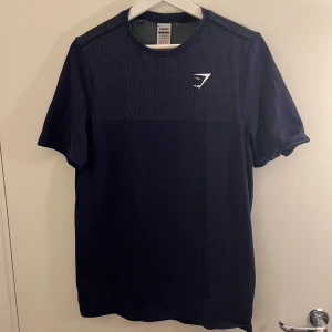 Mörkblå Gymshark t-shirt med mönster - Snygg mörkblå t-shirt från Gymshark i storlek S. Övre delen har ett diskret prickigt mönster och ett vitt Gymshark-logotryck på bröstet. T-shirten har rund hals och korta ärmar, perfekt för träning eller chill. Materialet är mjukt och känns lätt mot huden.
