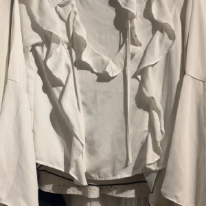 Vit blus med volanger och knyt - Supersnygg vit blus med volangdetaljer längs framstycket och knytband vid halsen. Blusen har långa ärmar med lätt utsvängd form och är gjord i ett mjukt, flowy material som ger en chill vibe. Perfekt för dig som gillar romantiska detaljer och vill sticka ut.