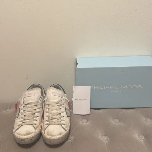 Philippe Model PRSX sneakers blå/vit - Säljer ett par Philippe Model PRSX Low sneakers i vitt skinn med blå och röda detaljer. Klassisk rund tå, snörning och platt sula. Märkeslogga på hälen och sidan, samt coola kontrastpaneler. Perfekt för dig som gillar fransk streetstyle och snygga detaljer.