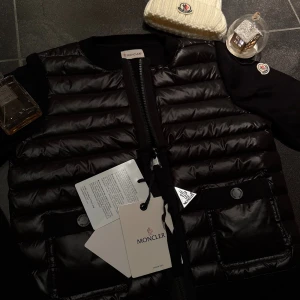 Svart cardigan från Moncler - Snygg svart cardigan från Moncler med quiltad design och två fickor framtill med texten ”moncler” skrivet på dem! Västen har en dragkedja och ribbad krage 😸. Materialet är glansigt och känns lyxigt, perfekt inför kallare dagar! Skickas självklart i originalförpackningen!😽💕