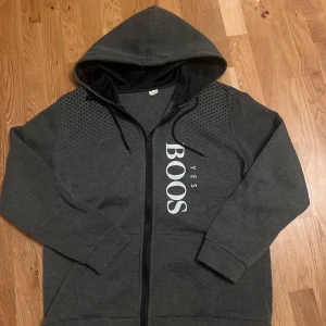 Mörkgrå hoodie med tryck YES BOOS - Mörkgrå hoodie från Mister Tee i storlek M med svart foder i huvan och trycket YES BOOS i vitt på framsidan. Tröjan har dragkedja, snörning i huvan och diskreta prickdetaljer på axlarna. Perfekt för chill eller streetwear.