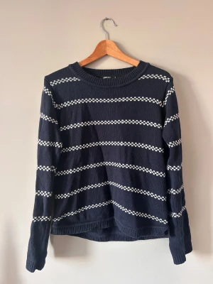 Marinblå randig stickad tröja M - Snygg marinblå stickad tröja från Serious Sally i storlek M. Tröjan har vita horisontella ränder med små mönster, rund hals och ribbade muddar vid ärmslut och nederkant. Perfekt för dig som gillar klassisk stil med en twist.