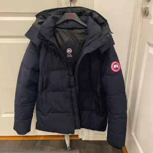 Mörkblå dunjacka från Canada Goose - Säljer en mörkblå dunjacka från Canada Goose med huva och klassisk logga på ärmen. Jackan har quiltad design, dragkedja och knappar framtill. Perfekt för kalla dagar och har en riktigt schysst passform. Fodret är i polyester och jackan är riktigt varm. Helt ny oanvänd