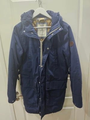 Blå parkas från Jack & Jones J&J75 - Mörkblå parkas från Jack & Jones J&J75 med huva, dragkedja och knappar framtill. Jackan har flera fickor med lock och knappar samt quiltat, guldigt innerfoder. Perfekt för kyliga dagar och snygg streetstil.