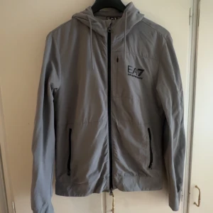 Grå vindjacka från EA7 Emporio Armani - Snygg grå vindjacka från EA7 Emporio Armani med huva och svarta dragkedjor. Jackan har logga på bröstet, två fickor med dragkedja och justerbar snörning i huvan. Perfekt för dig som gillar stilren och sportig design.
