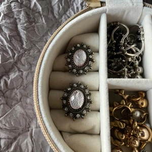 Vintageinspirerade örhängen med stenar - Säljer ett par stora, ovala örhängen med antikguldig metall och ljusrosa, mönstrad mittsten. Runt stenen sitter små klara stenar i en blomformad design. Perfekta för dig som gillar statement-smycken med retrovibe.