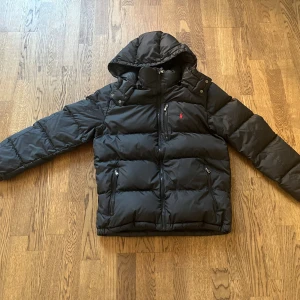 Ralph Lauren vinterjacka - Svart pufferjacka från Polo Ralph Lauren med huva och dragkedja. Jackan har två sidofickor med dragkedja och en bröstficka, samt den klassiska röda Polo-loggan broderad på bröstet. Perfekt för kalla dagar och riktigt snygg streetstil. Stl XL (18-20) motsvarar stl XS. 